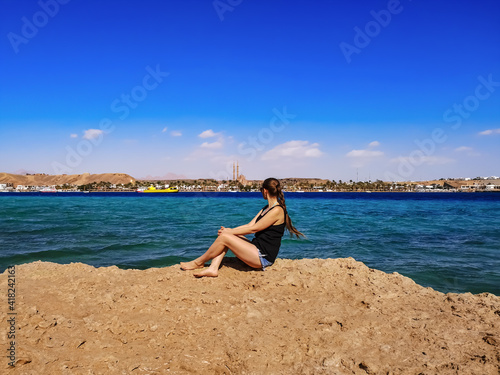 Barefoot young woman with blond braided hair sits on a rock against the background of the sea panorama of Sharm El Sheikh (Egypt). Female tourist looks out over the Sharm El Maya bay © ioanna_alexa