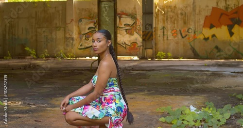 Sexy young girl in a long flower dress turns while squating at an abandoned warehouse on the Caribbean island of Trinidad photo