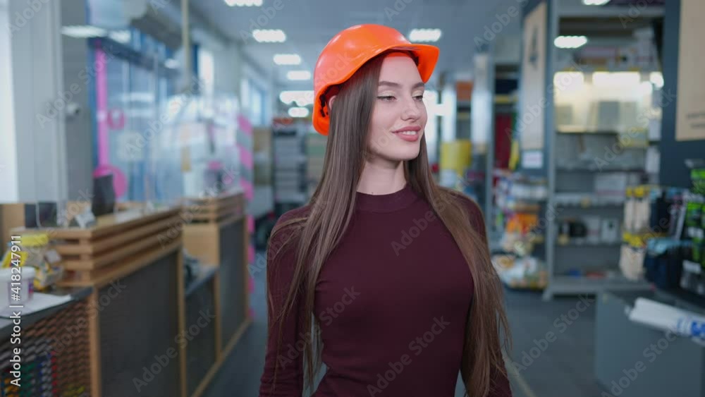 Portrait of elegant slim beautiful woman in hard hat posing in hardware store indoors. Charming brunette Caucasian buyer with brown eyes looking at camera smiling standing between rows with goods.