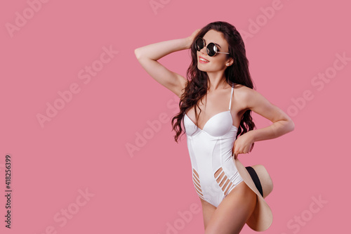 Charming girl in white one-piece swimsuit and straw hat isolated over pink background.