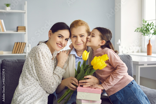Portrait of three generations of women, mom, grandma and granddaughter celebrating event. Girl with mother congratulating grandmother on birthday, March 8 or Mother's Day handing gifts and flowers.