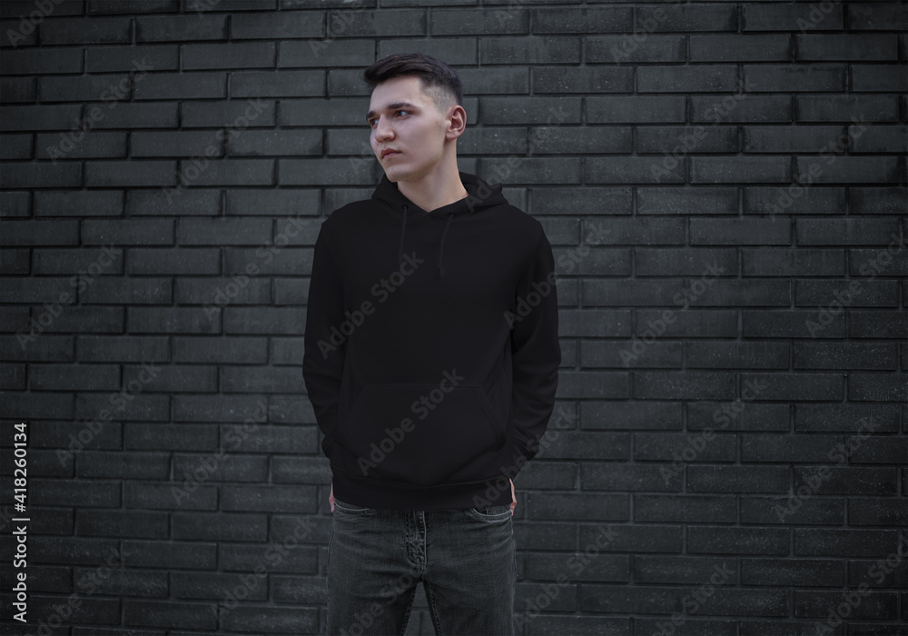 Black template with a hood on a young guy with hands in pockets, blank mens clothing on a brick wall background, front view. Black template with a hood on a young guy with hands in pockets, blank mens clothing on a brick wall background, front view.