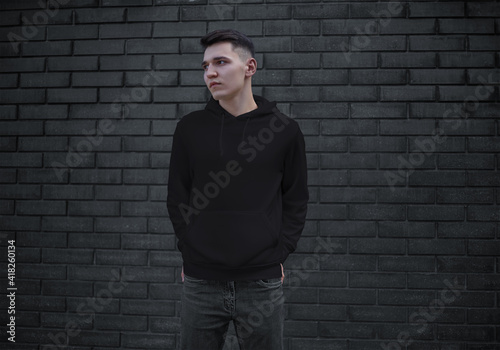 Black template with a hood on a young guy with hands in pockets, blank mens clothing on a brick wall background, front view.