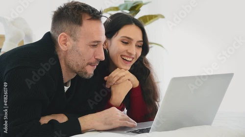 Man and woman couple in casual winter outfit lay down on bed using laptop with happy and romantic mood in bedroom