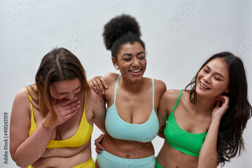 Three joyful diverse young women with different body shapes wearing colorful underwear laughing, posing together over light background