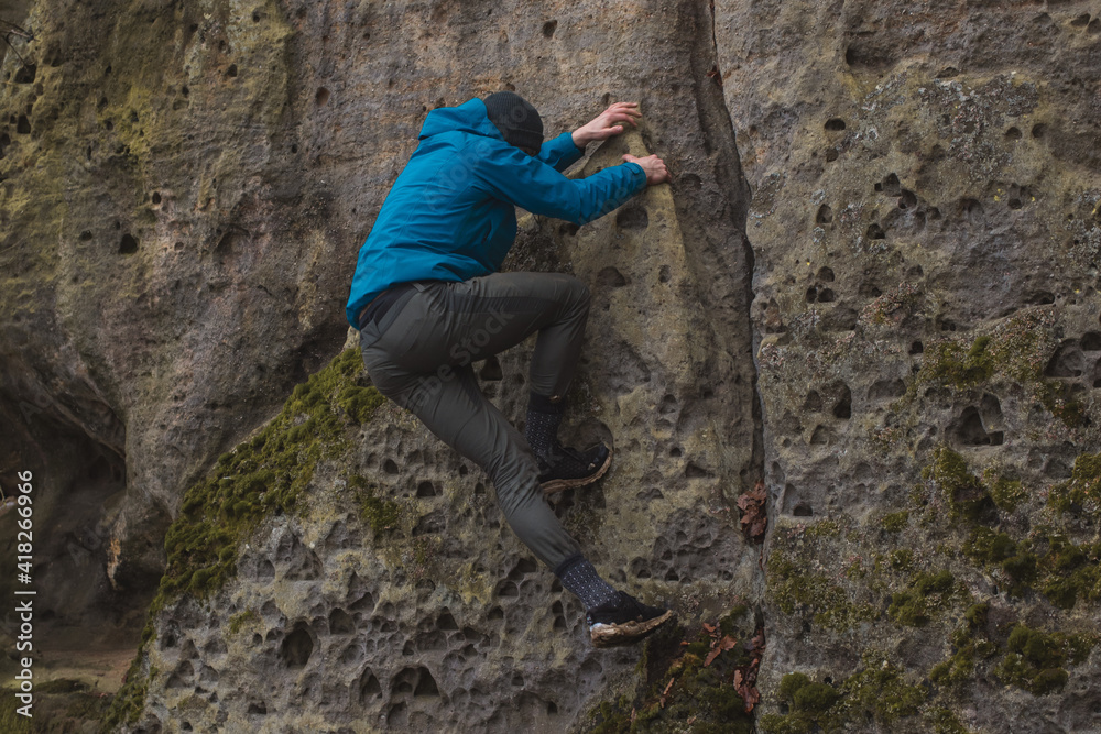 man in a blue jacket climbing a stone wall holding with strong hands in a crack in the stone wall
