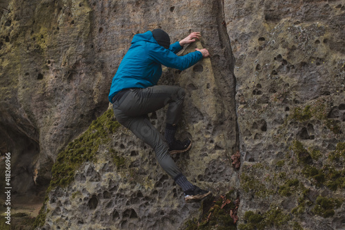 man in a blue jacket climbing a stone wall holding with strong hands in a crack in the stone wall