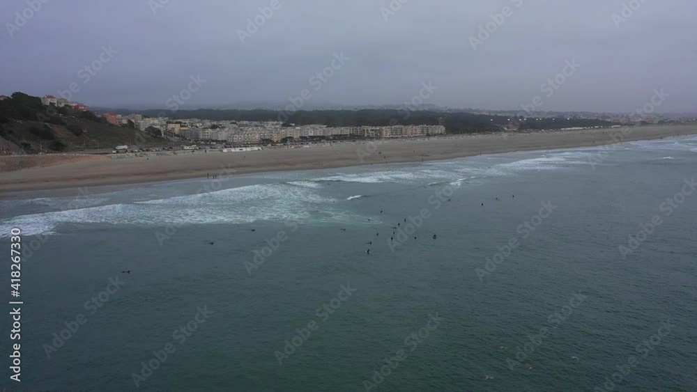 Aerial shot of surfers on waves at beach against sky, drone descending forward towards coastline at sunset - San Francisco, California