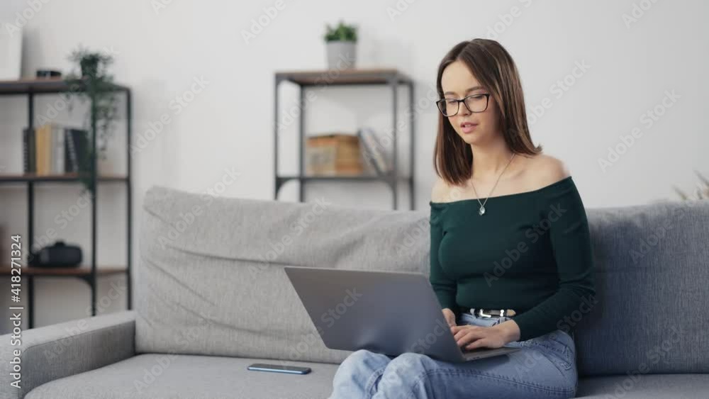 Happy young lady in eyeglasses surfing internet on wireless laptop while sitting on cozy sofa. Pretty woman in stylish outfit staying at home and using modern gadget. Happy young lady in eyeglasses surfing internet on wireless laptop while sitting on cozy sofa. Pretty woman in stylish outfit staying at home and using modern gadget.