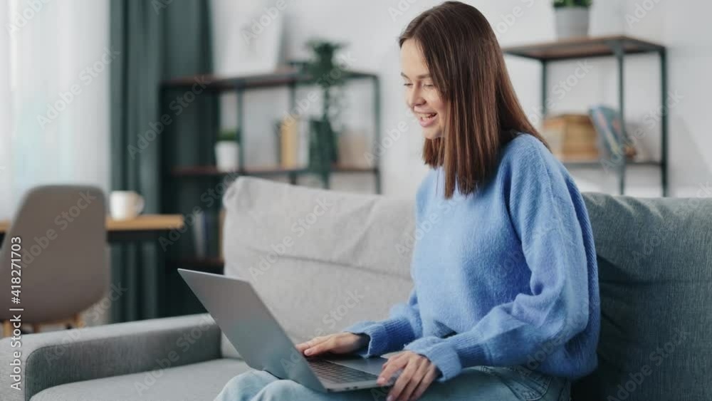 Positive young woman in blue sweater and jeans talking and gesturing during video call on wireless laptop. Happy brunette having online conversation while sitting on couch at home.