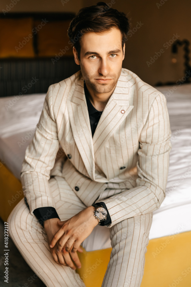 Handsome respectable groom is sitting on the bed of a luxury hotel room, the groom's light wedding suit