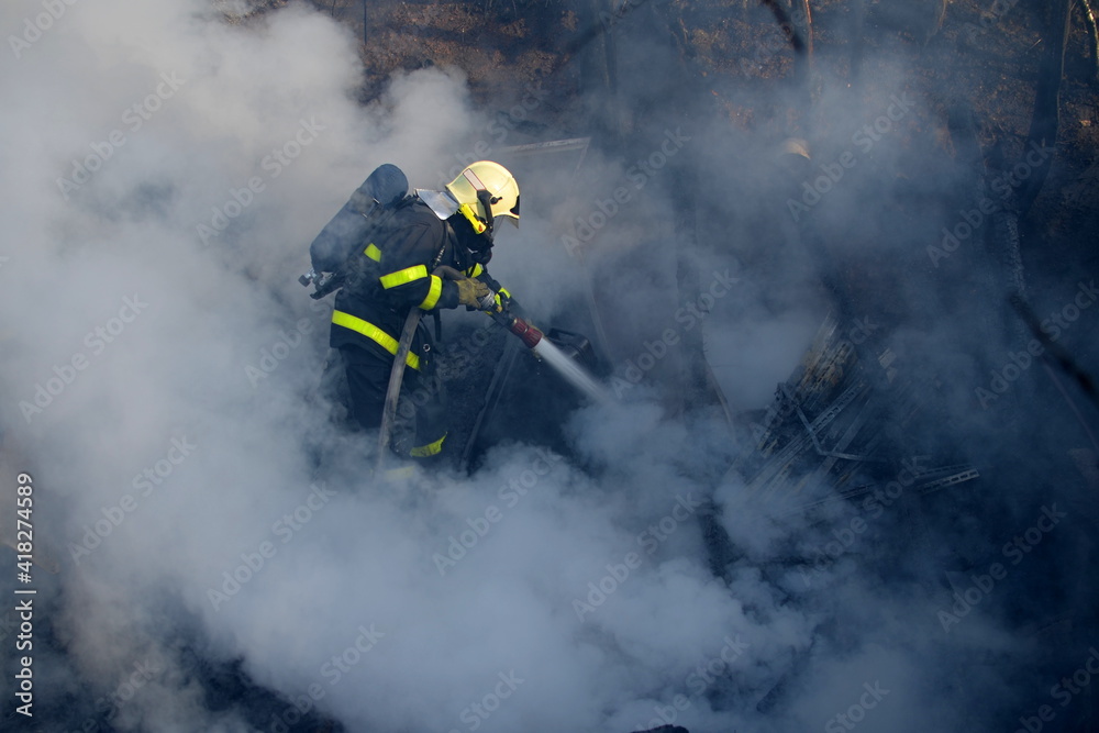 Fototapeta premium Firefighter with a breathing apparatus shrouded and surrounded by thick smoke extinguish the fire of wooden buildings with water from a hose