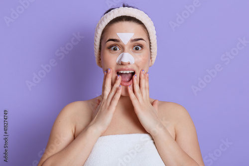 Excited lady posing with white skin stickers on nose and forehead, isolated over lilac background, doing cleansing procedures, keeping mouth opened, touching her cheeks, being surprised and amazed.