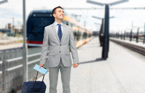 business trip and people concept - happy businessman in suit with travel bag and ticket over train on railway station on background