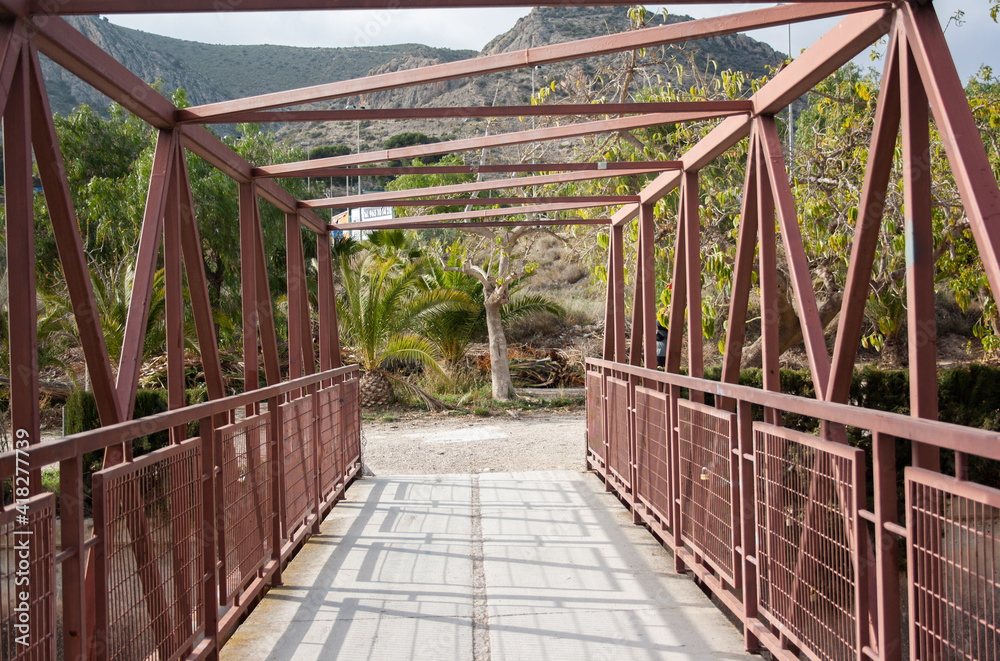 Red metal bridge over the Vinalopo river in a public park in Elda, Alicante, with Bolon mountain in the background.
