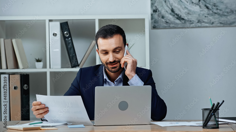 Smiling businessman talking on smartphone while working with papers in office Smiling businessman talking on smartphone while working with papers in office