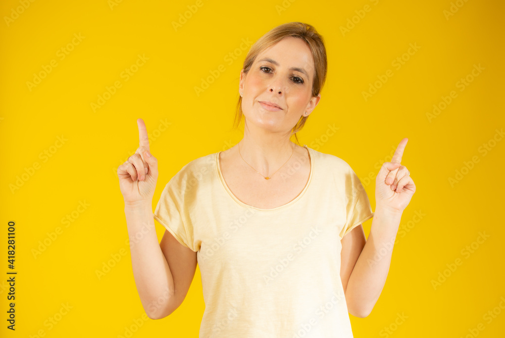 Portrait of a young woman dressed in casual t-shirt pointing fingers up at copy space isolated over yellow background Portrait of a young woman dressed in casual t-shirt pointing fingers up at copy space isolated over yellow background