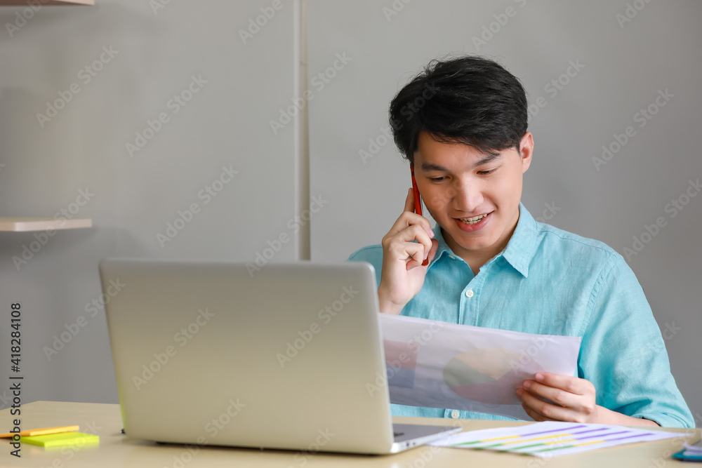 Young handsome male businessman with black short hair wearing light blue shirt working on office desk with silver laptop by looking at work document while using red mobile phone