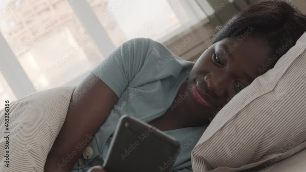 Close up of young beautiful African woman wearing pajamas lying in bed at home and using her smartphone Close up of young beautiful African woman wearing pajamas lying in bed at home and using her smartphone