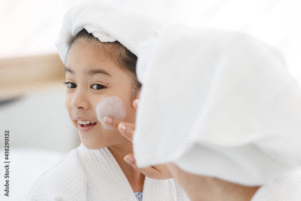 Mother adding treatment cream on the cheek to young and cute Asian girl with spa dress and head covered with a white towel. The kid's face expresses a happy smile and joy Mother adding treatment cream on the cheek to young and cute Asian girl with spa dress and head covered with a white towel. The kid's face expresses a happy smile and joy