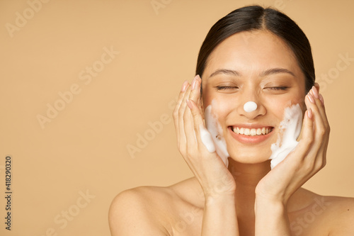 Studio portrait of pleased young woman smiling with eyes closed while applying gentle foam facial cleanser isolated over beige background