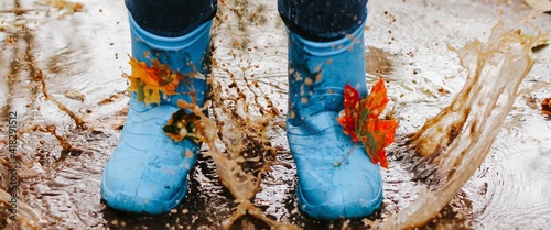 Teenager girl wearing blue rain boots jumping into a puddle on rainy autumn day. Closeup. Banner.	