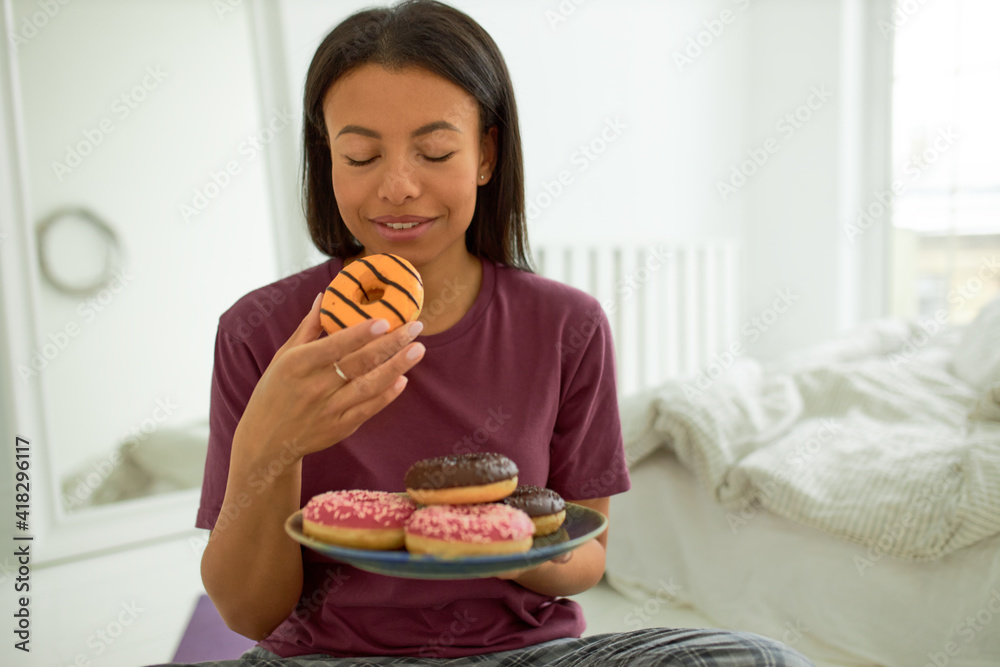 Indoor image of attractive young mixed race woman in pajamas sitting in bedroom enjoying happy moment of pleasure and satisfaction holding big glazed delicious doughnut, closing eyes, going to bite Indoor image of attractive young mixed race woman in pajamas sitting in bedroom enjoying happy moment of pleasure and satisfaction holding big glazed delicious doughnut, closing eyes, going to bite