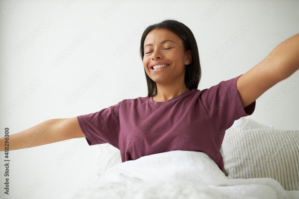 Happy young dark skinned female in pajamas sitting on white bed with arms outstretched, keeping eyes closed, smiling broadly, being grateful for new day, stretching body, full of energy and joy Happy young dark skinned female in pajamas sitting on white bed with arms outstretched, keeping eyes closed, smiling broadly, being grateful for new day, stretching body, full of energy and joy