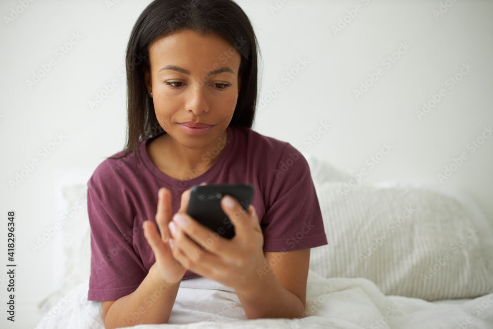 Attractive stylish young Afro American female in cozy t-shirt sitting on bed holding mobile, checking email in the morning right after awakening. Pretty girl surfing internet on smart phone in bedroom Attractive stylish young Afro American female in cozy t-shirt sitting on bed holding mobile, checking email in the morning right after awakening. Pretty girl surfing internet on smart phone in bedroom