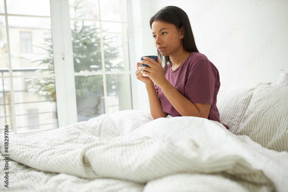 People, beverage, relaxation and coziness. Good looking young dark skinned woman sitting in bed early in the morning holding cup, having coffee or hot chocolate, planning day, enjoying favorite drink People, beverage, relaxation and coziness. Good looking young dark skinned woman sitting in bed early in the morning holding cup, having coffee or hot chocolate, planning day, enjoying favorite drink