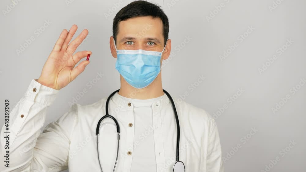 A young handsome doctor in a medical mask holds a red pill in his hands presenting a new drug.A young doctor shows a new drug at the presentation, a red pill for all diseases. A young handsome doctor in a medical mask holds a red pill in his hands presenting a new drug.A young doctor shows a new drug at the presentation, a red pill for all diseases.