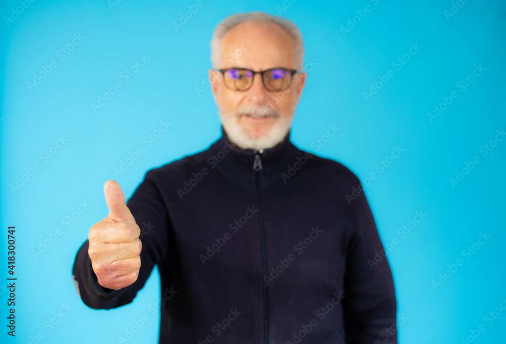 Senior grey-haired man standing over isolated blue background doing happy thumb up gesture with hand. Approving expression looking at the camera showing success. Senior grey-haired man standing over isolated blue background doing happy thumb up gesture with hand. Approving expression looking at the camera showing success.
