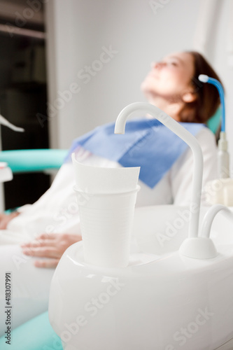 detail of dental chair with glass of water and tap, with a brunette female patient, in the background, © Fernando