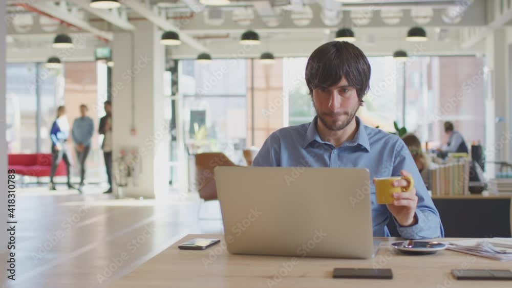 Dolly shot of casually dressed businessman working on laptop taking coffee break at desk in modern open plan start up office with colleagues in background - shot in slow motion Dolly shot of casually dressed businessman working on laptop taking coffee break at desk in modern open plan start up office with colleagues in background - shot in slow motion