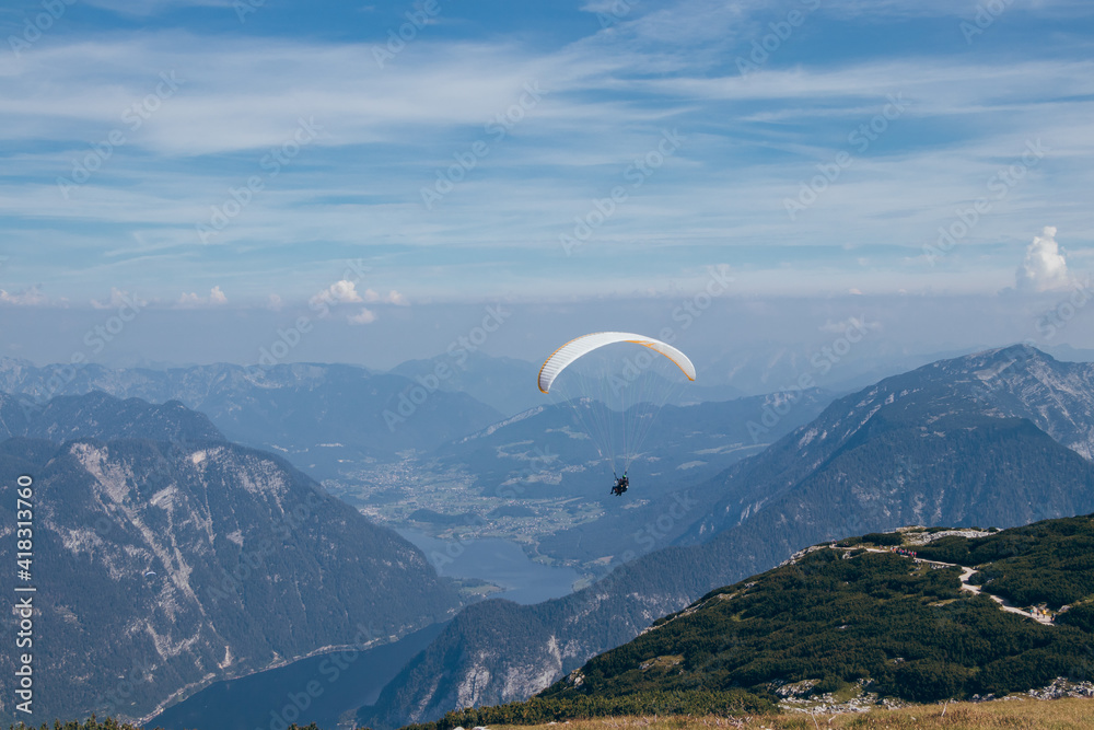 Man with white-and-yellow parachute hovers over Mount Krippenstein in upper Austria, enjoying view of valley and rest of the Austrian alps. Ideal sunny weather without gusty winds for paragliding Man with white-and-yellow parachute hovers over Mount Krippenstein in upper Austria, enjoying view of valley and rest of the Austrian alps. Ideal sunny weather without gusty winds for paragliding