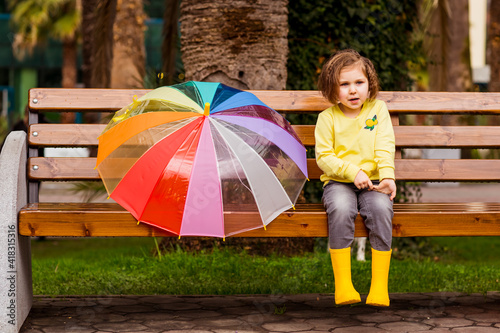 a beautiful little girl in yellow boots and a yellow sweater is sitting on a park bench with a colored umbrella