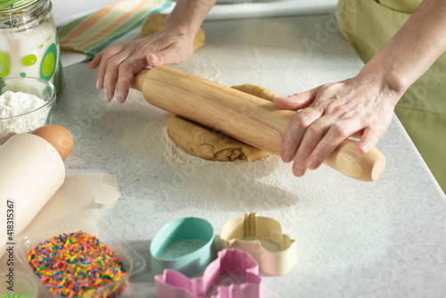 Female model rolls dough with rolling pin on kitchen table. Cookie cutters and sugar sprinkles aside.