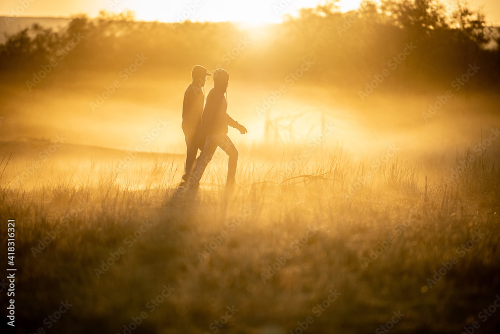 Two people walking through the grass one early morning in winter with the sun coming up in the distance.