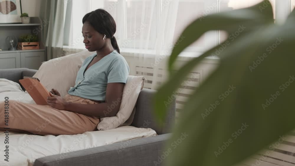 Medium shot of young African-American woman wearing wireless earphones sitting in bed at home with digital tablet in hands, messaging with friend and listening to music Medium shot of young African-American woman wearing wireless earphones sitting in bed at home with digital tablet in hands, messaging with friend and listening to music