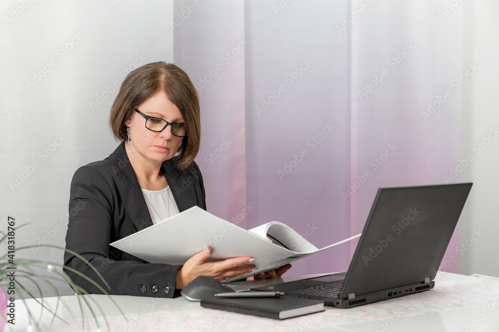 A brunette bussiness woman in glasses in a black jacket works on the computer in the office holds a folder with documents in his hands and reads A brunette bussiness woman in glasses in a black jacket works on the computer in the office holds a folder with documents in his hands and reads
