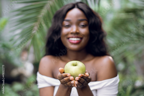 People, beauty, nature and healthy fruit. Blurred front close up view of young cheerful African woman in white t-shirt, posing with smile to camera, holding green apple, on exotic plants background. © sofiko14