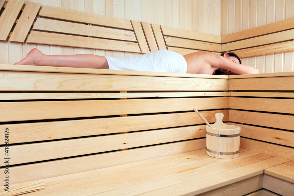 Slim woman wrapped in white towel lies on the bench in a traditional sauna