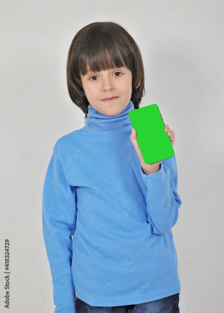 Mobile app for children. Happy excited cheerful little caucasian boy with dark hair pointing at cell phone and smiling at camera, showing telephone. indoor studio shot isolated on grey background Mobile app for children. Happy excited cheerful little caucasian boy with dark hair pointing at cell phone and smiling at camera, showing telephone. indoor studio shot isolated on grey background