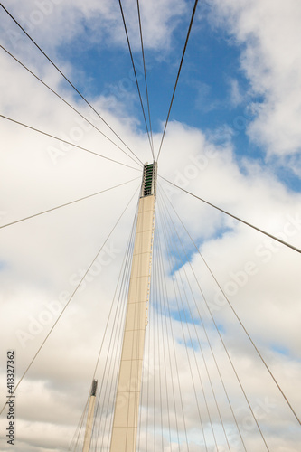 Architecture detail of a Pedestrian bridge support tower and cables against a blue sky with puffy clouds