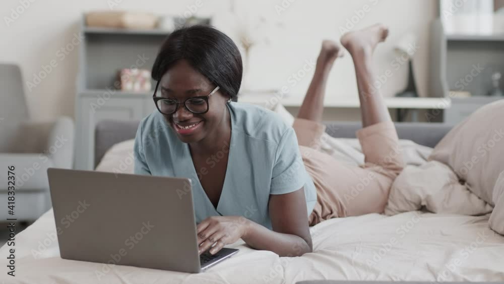 Lockdown of young joyful African-American woman wearing comfortable clothes and eyeglasses lying in bed at home and messaging with friend using laptop Lockdown of young joyful African-American woman wearing comfortable clothes and eyeglasses lying in bed at home and messaging with friend using laptop