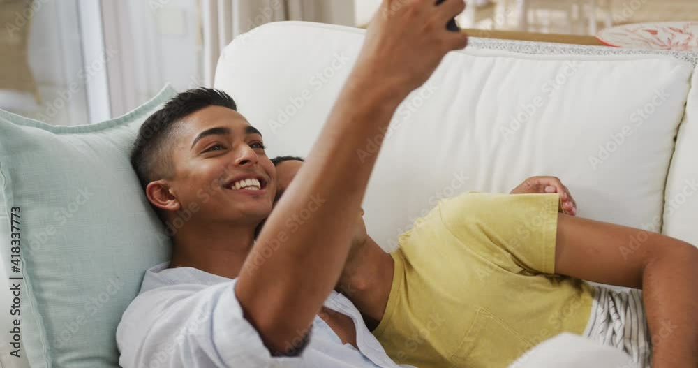 Smiling mixed race gay male couple hugging lying on sofa taking selfie Smiling mixed race gay male couple hugging lying on sofa taking selfie