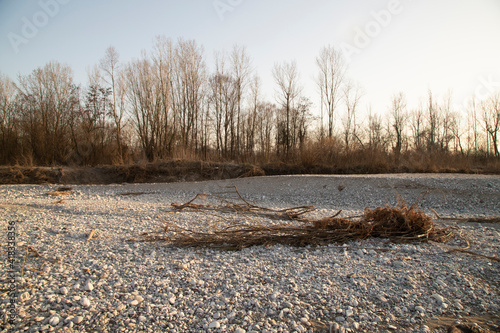 River scenery with grey cobblestone and river debris with trees and bushes in the background.
