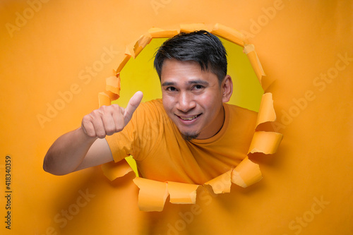 Young entertaining man through torn yellow paper hole showing thump up with facetious of content and happiness