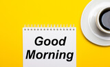 On a bright yellow background, a white cup with coffee and a white notepad with the words GOOD MORNING