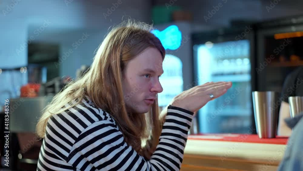 Young Caucasian man with long hair talking and gesturing sitting at bar counter in pub. Portrait of relaxed millennial resting indoors chatting with friend. Relaxation and lifestyle. Young Caucasian man with long hair talking and gesturing sitting at bar counter in pub. Portrait of relaxed millennial resting indoors chatting with friend. Relaxation and lifestyle.